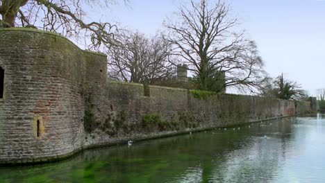 Over-the-moat,-looking-towards-the-imposing-walls-and-castle-towers-of-the-Medieval-Bishop's-Palace-in-Wells,-UK