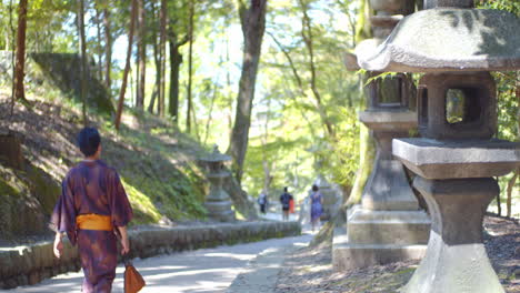 Old-stone-lantern,-guy-wearing-a-Yukata-walking-dow-a-path-in-the-woods-in-Kyoto,-Japan-soft-lighting