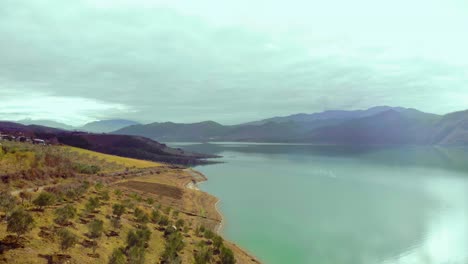 New-trees-planted-on-shore-of-mountain-lake,-vivid-colors-of-sunny-winter-day-with-grey-clouds,-drone-footage