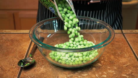 Cook-pours-frozen-peas-from-plastic-packaging-into-glass-bowl,-slowmo