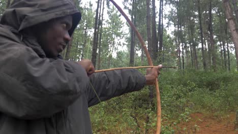 A-slow-motion-orbit-shot-around-a-hooded-African-hunter-shooting-a-traditional-wooden-bow-and-arrow-in-a-forest