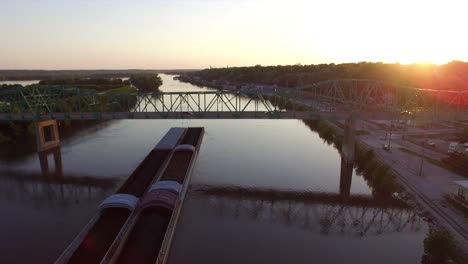 Cargo-boat-transportation-slowly-driving-under-a-bridge-in-canal-during-sunset,-Aerial