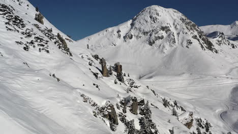 Drone-shot,-dolly-forward-along-a-snow-covered-mountain-side-with-rock-pinnacles,-towards-a-large-mountain,-with-ski-runs-visible-in-La-Plagne-France