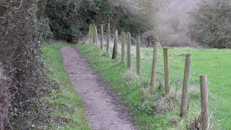 A-footpath-alongside-a-thick-hedgerow-and-some-fenceposts-supporting-barbed-wire