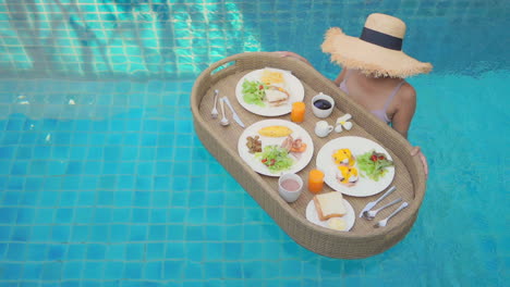 Female-tourist-preparing-to-eat-a-breakfast-served-on-a-floating-tray-in-the-swimming-pool-in-a-luxurious-tropical-hotel