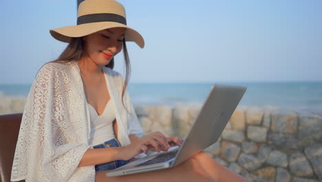 Close-up-of-a-young-Asian-woman-typing-on-her-laptop-as-she-enjoys-the-ocean-breezes