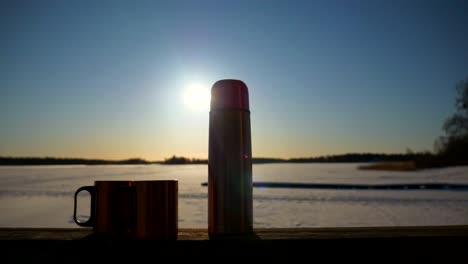 Close-up-View-of-cup-of-hot-coffee-and-thermos,-Sun-and-snow-background