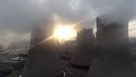 Aerial-view-of-power-station,-sunrise-moving-behind-cooling-towers-smoke-steam-emissions