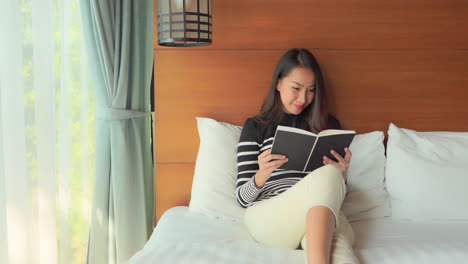 Asiatische-Frau-Mit-Einem-Schönen-Lächeln-Liest-Eine-Zeitschrift,-Die-Auf-Einem-Weißen-Hotelzimmerbett-Neben-Dem-Fenster-Sitzt