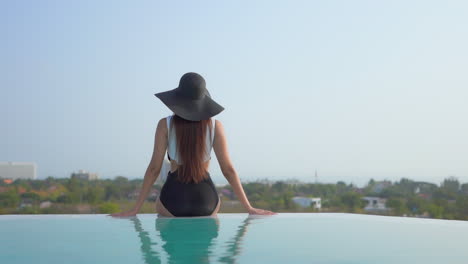 A-woman-perched-on-the-edge-of-a-resort-pool-sits-with-her-back-to-the-camera-as-she-looks-over-the-cityscape-beyond