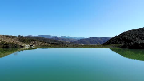 Lake-surface-like-glassy-mirror-reflecting-blue-sky,-bordered-by-reservoir-dam-on-top-of-mountains,-peaceful-Alpine-panorama