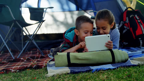 Siblings-using-digital-tablet-outside-the-tent-at-campsite
