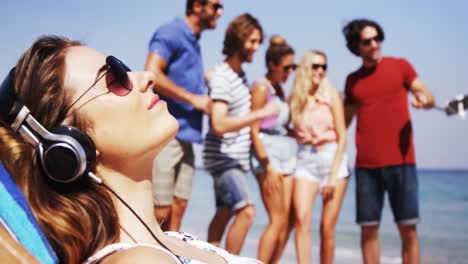 Beautiful-woman-listening-to-music-while-friends-taking-a-selfie-at-beach