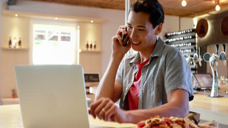 Mann-Benutzt-Laptop,-Während-Er-In-Einem-Restaurant-Mit-Dem-Mobiltelefon-Telefoniert-4k