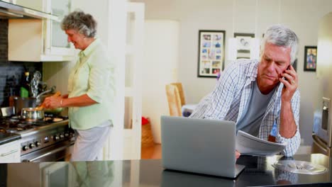 Älterer-Mann-Benutzt-Laptop,-Während-Frau-Im-Hintergrund-4k-Kocht