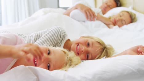 Lächelnde-Kinder-Entspannen-Sich-Auf-Dem-Bett,-Während-Die-Eltern-Im-Hintergrund-Schlafen,-4k