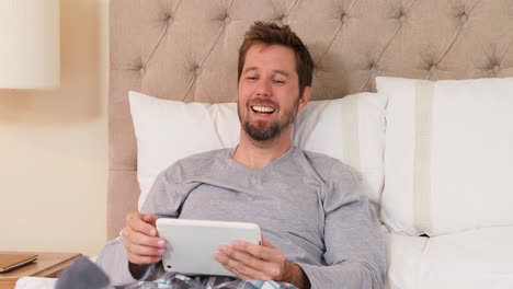 Smiling-young-man-sitting-on-bed-using-his-tablet-in-the-bedroom-4K-4k