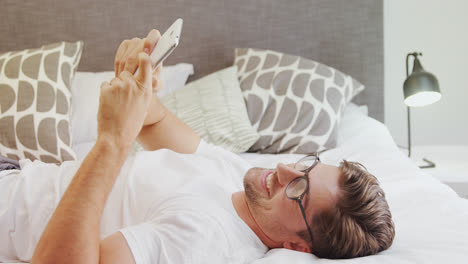 Smiling-man-wearing-spectacles-lying-on-bed-using-mobile-phone-4K-4k