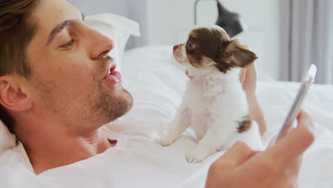 Young-man-lying-on-bed-stroking-his-puppy-while-using-mobile-phone-4K-4k