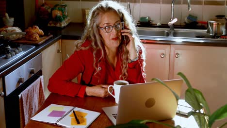 Mature-woman-having-coffee-while-talking-on-mobile-phone-in-kitchen-4k