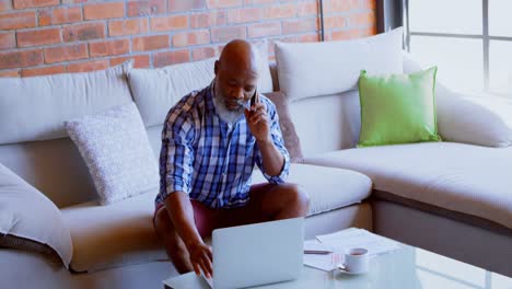 Senior-man-using-laptop-while-talking-om-mobile-phone-in-living-room-4k
