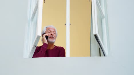 Älterer-Mann,-Der-Zu-Hause-Am-Fenster-Mit-Dem-Mobiltelefon-Telefoniert,-4K