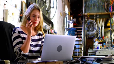 Woman-talking-on-mobile-phone-while-using-laptop-at-workshop-4k