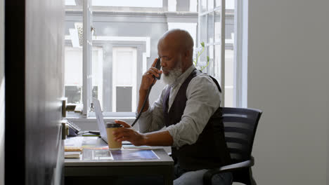 Businessman-having-coffee-while-talking-on-landline-and-using-laptop-at-desk-4k