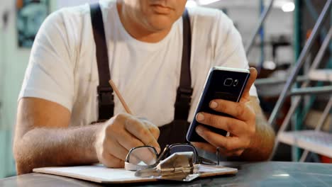 Male-mechanic-writing-on-clipboard-while-using-mobile-phone-4k
