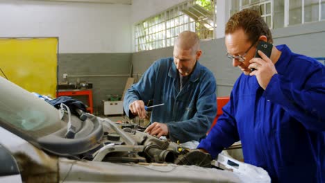 Mechanic-examining-a-car-while-his-colleague-talking-on-mobile-phone-4k