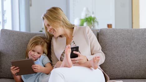 Mother-and-daughter-using-mobile-phone-and-digital-tablet-in-living-room-4k