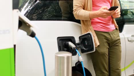 Man-using-mobile-phone-while-charging-electric-car-at-charging-station-4k