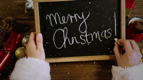 Video-composition-with-falling-snow-over-desk-with-santa-holding-blackboard-with-merry-Christmas