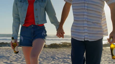 Mid-section-of-couple-holding-hands-with-beer-bottle-walking-on-beach-4k