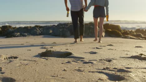 Couple-holding-hands-with-beer-bottle-walking-on-beach-4k