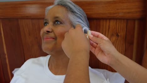 Female-doctor-inserting-hearing-aid-in-senior-black-woman-ear-in-a-comfortable-home-4k