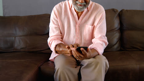 Vorderansicht-Eines-älteren-Schwarzen-Mannes,-Der-Auf-Der-Couch-Sitzt-Und-Sein-Mobiltelefon-In-Einem-Komfortablen-Zuhause-Benutzt,-4K