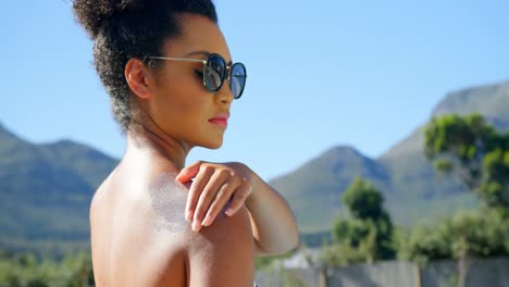 Side-view-of-young-mixed-race-woman-applying-sunscreen-on-her-shoulder-near-swimming-pool-4k