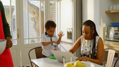 Front-view-of-black-young-mother-working-on-laptop-and-son-playing-at-dining-table-in-kitchen-4k