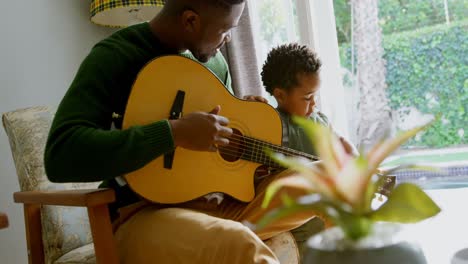 -Side-view-of-young-black-father-and-little-son-playing-guitar-in-living-room-of-comfortable-home-4k