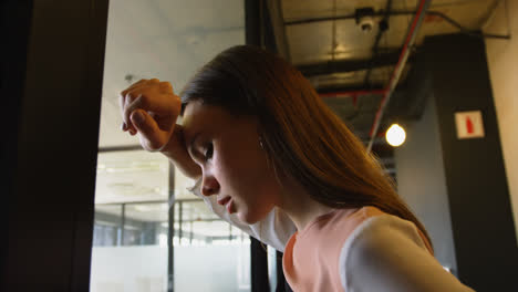Low-angle-side-view-of-young-Caucasian-businesswoman-leaning-head-against-glass-wall-in-modern-offic