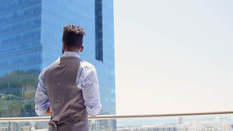 Side-view-of-young-black-businessman-texting-on-mobile-phone-and-standing-in-office-balcony-4k