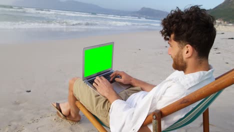 Side-view-of-young-caucasian-man-sitting-on-lounger-and-using-laptop-at-beach-4k