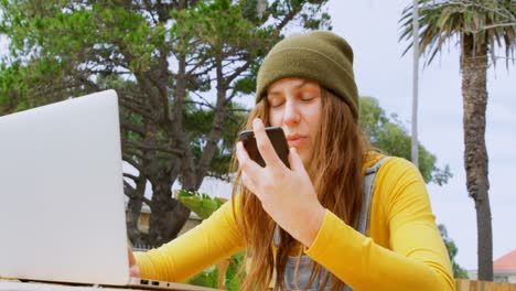 Young-caucasian-female-skateboarder-talking-on-mobile-phone-while-using-laptop-at-outdoors-cafe-4k