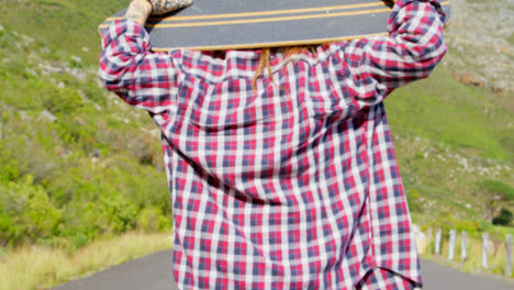 Rear-view-of-cool-young-female-skateboarder-carrying-skateboard-on-shoulder-at-countryside-road-4k