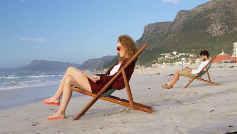 Side-view-of-young-caucasian-woman-with-sunglasses-sitting-on-lounger-and-reading-a-book-at-beach-4k