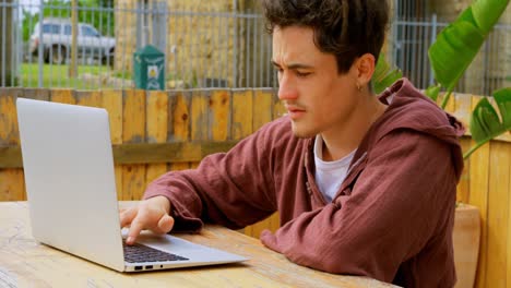 Side-view-of-cool-young-caucasian-male-skateboarder-working-on-laptop-at-outdoor-cafe-4k