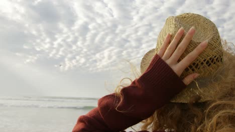 Rear-view-of-young-blonde-woman-holding-hat-and-looking-at-sea-on-the-beach-4k