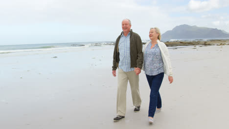 Front-view-of-old-caucasian-couple-walking-hand-in-hand-at-beach-4k