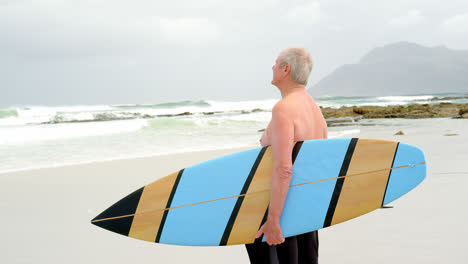 Side-view-of-old-caucasian-man-standing-with-surfboard-at-beach-4k